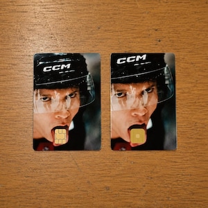 Op de afbeelding: Twee rechthoekige kaarten met het gezicht van een hockeyspeler, die een zwarte helm met wit "CCM" draagt. De mond van de speler is open, met een klein, vierkant, goudkleurig object op de tong.
