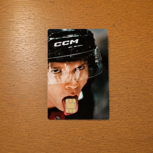 Op de afbeelding: Een rechthoekige kaart met een hockey speler met een zwarte helm en het woord "CCM". Het gezicht van de speler is zichtbaar, met een simkaart in plaats van de tong. De achtergrond is een houten oppervlak.