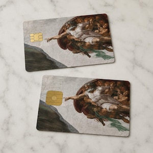 May include: Two rectangular cards featuring a section of Michelangelo's "The Creation of Adam" painting. The cards have a gold-colored chip on the left side. The artwork depicts two figures with outstretched arms, nearly touching.