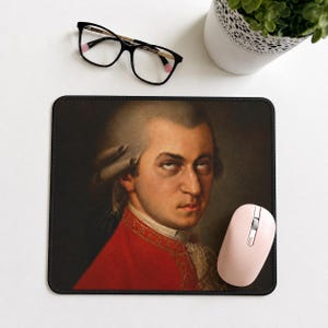 May include: A rectangular mouse pad featuring a portrait of Wolfgang Amadeus Mozart wearing a red coat with gold embroidery and a white cravat. The image shows the composer with a powdered wig, looking directly forward against a dark background.
