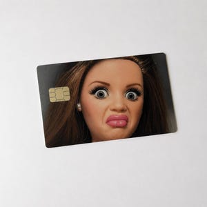 Op de afbeelding: Een rechthoekige creditcard met een poppengezicht erop gedrukt. De pop heeft bruin haar, grote ogen en een verbaasde uitdrukking. Een goudkleurige chip bevindt zich in de linkerbovenhoek van de kaart.