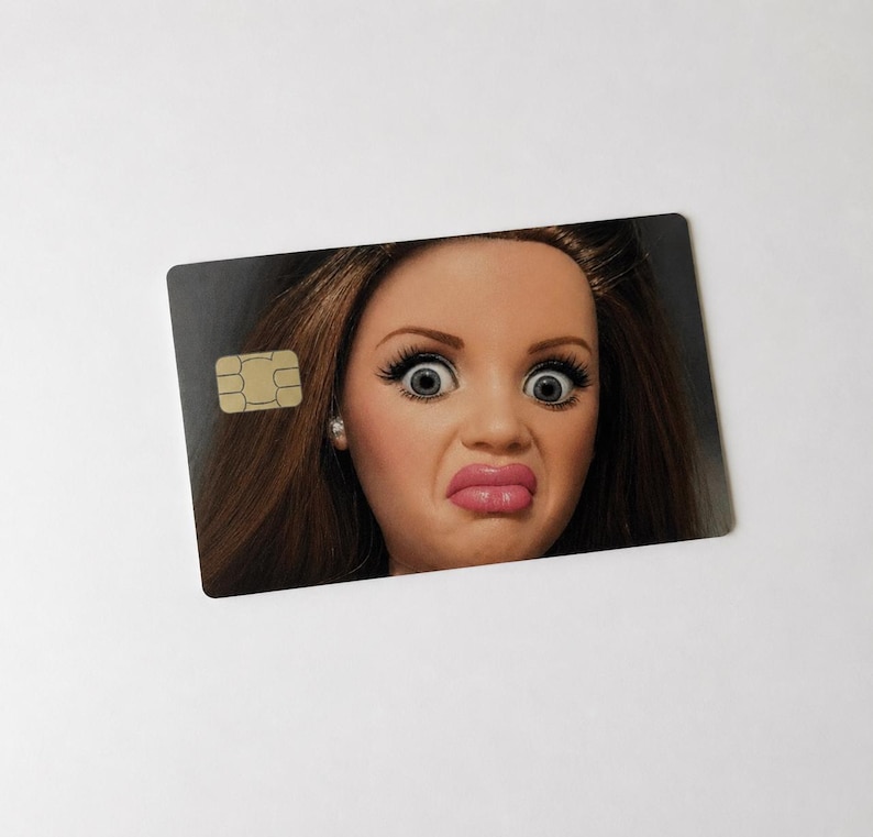 Op de afbeelding: Een rechthoekige creditcard met een poppengezicht erop gedrukt. De pop heeft bruin haar, grote ogen en een verbaasde uitdrukking. Een goudkleurige chip bevindt zich in de linkerbovenhoek van de kaart.