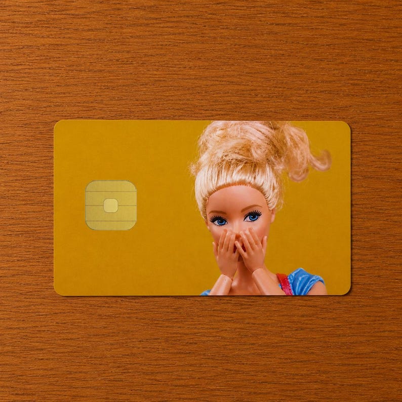 Pu&ograve; includere: Una carta di pagamento rettangolare con sfondo giallo senape che presenta l'immagine stampata di una bambola bionda con occhi azzurri e capelli raccolti in uno chignon spettinato, che si copre la bocca con entrambe le mani. Chip dorato a sinistra.
