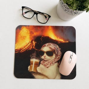 Peut inclure: Tapis de souris carré avec un chat blanc portant des lunettes de soleil et un foulard, tenant une boisson au café, avec un volcan en arrière-plan. Une souris d'ordinateur rose et des lunettes noires sont également visibles.