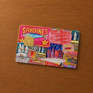 May include: A decorative credit card featuring a colorful sardine design. The card has a pink background with illustrations of sardines, sardine cans, and the word "Sardines" in large letters. The card is on a wooden surface.