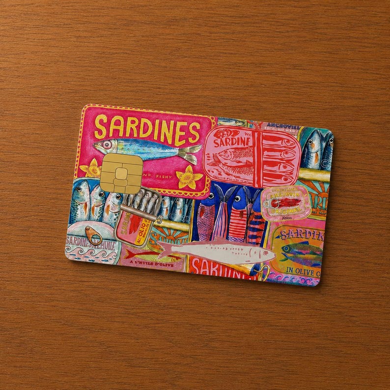 May include: A decorative credit card featuring a colorful sardine design. The card has a pink background with illustrations of sardines, sardine cans, and the word "Sardines" in large letters. The card is on a wooden surface.