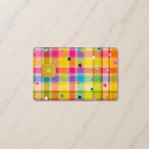 Peut inclure: Une carte de cr&eacute;dit rectangulaire pr&eacute;sentant un motif &agrave; carreaux multicolore vif dans des tons de jaune, rose, orange et bleu. La surface est d&eacute;cor&eacute;e de nombreuses petites &eacute;toiles dispers&eacute;es de diff&eacute;rentes couleurs et d'une puce EMV dor&eacute;e.
