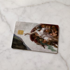 May include: A rectangular credit card featuring a reproduction of Michelangelo's "The Creation of Adam" fresco. The card has a gold-colored EMV chip on the left side. The artwork depicts God reaching out to Adam.