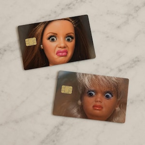 Op de afbeelding: Twee rechthoekige kaarten met afbeeldingen van poppengezichten met overdreven uitdrukkingen. De kaarten hebben een goudkleurige chip aan de linkerkant. E&eacute;n pop heeft bruin haar, de andere blond. Lichte marmeren achtergrond.