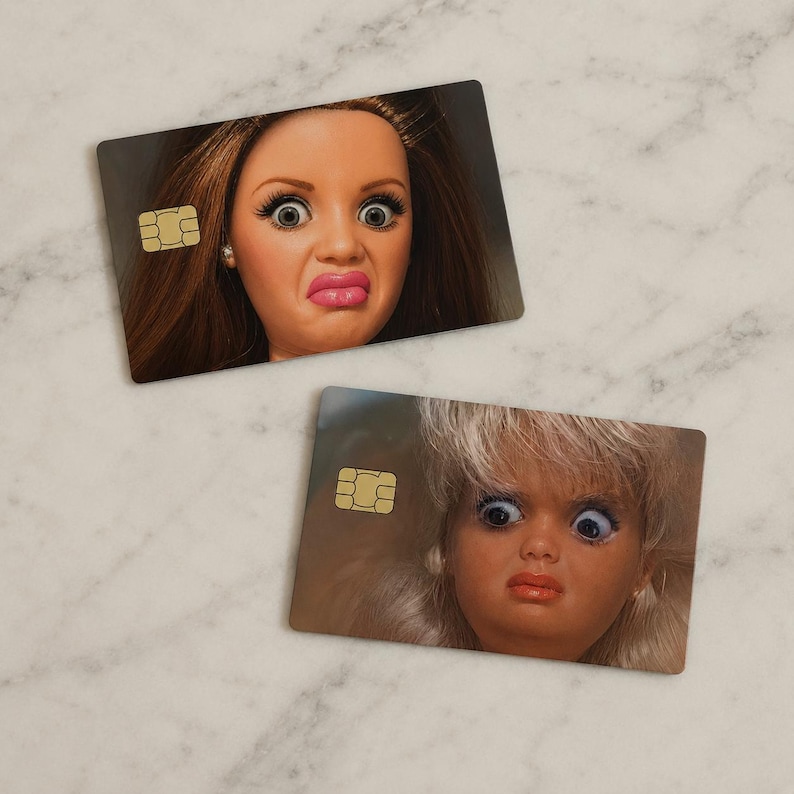 Op de afbeelding: Twee rechthoekige kaarten met afbeeldingen van poppengezichten met overdreven uitdrukkingen. De kaarten hebben een goudkleurige chip aan de linkerkant. E&eacute;n pop heeft bruin haar, de andere blond. Lichte marmeren achtergrond.