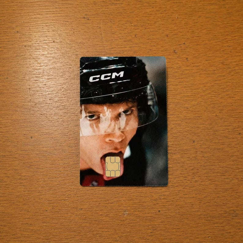 Op de afbeelding: Een rechthoekige kaart met een hockey speler met een zwarte helm en het woord "CCM". Het gezicht van de speler is zichtbaar, met een simkaart in plaats van de tong. De achtergrond is een houten oppervlak.