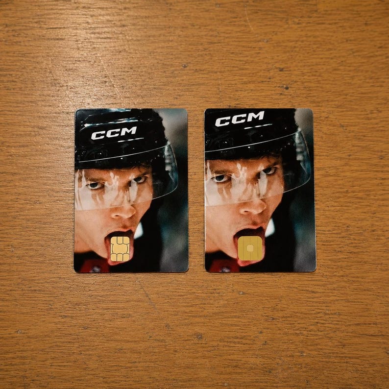 Op de afbeelding: Twee rechthoekige kaarten met het gezicht van een hockeyspeler, die een zwarte helm met wit "CCM" draagt. De mond van de speler is open, met een klein, vierkant, goudkleurig object op de tong.