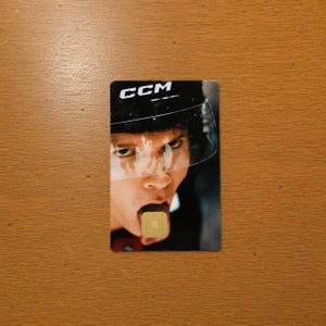 Op de afbeelding: Een rechthoekige kaart met een hockeyspeler met een zwarte helm en het "CCM"-logo. Het gezicht van de speler is zichtbaar, met zijn tong uit en een goudkleurige chip erop. De achtergrond is een houten oppervlak.