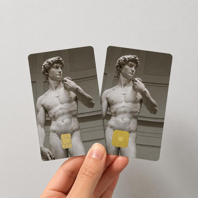 Op de afbeelding: Twee rechthoekige kaarten met een grijswaardenafbeelding van Michelangelo's David-standbeeld, met een goudkleurige chip die de geslachtsdelen van het standbeeld bedekt. De kaarten worden door een hand tegen een witte achtergrond gehouden.