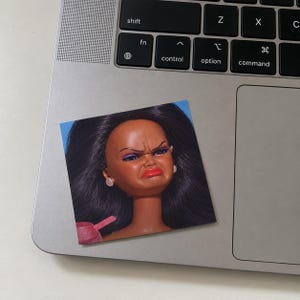 May include: A square sticker featuring a doll with long black hair, dark skin, and a facial expression of disgust. The doll wears small white stud earrings and has bright red lipstick. The sticker is placed on the metallic surface of a laptop.