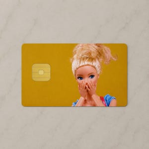 Pu&ograve; includere: Una carta di credito rettangolare con sfondo giallo senape, un chip EMV dorato sul lato sinistro e una bambola con capelli biondi raccolti in uno chignon, occhi azzurri e mani che coprono la bocca sul lato destro.