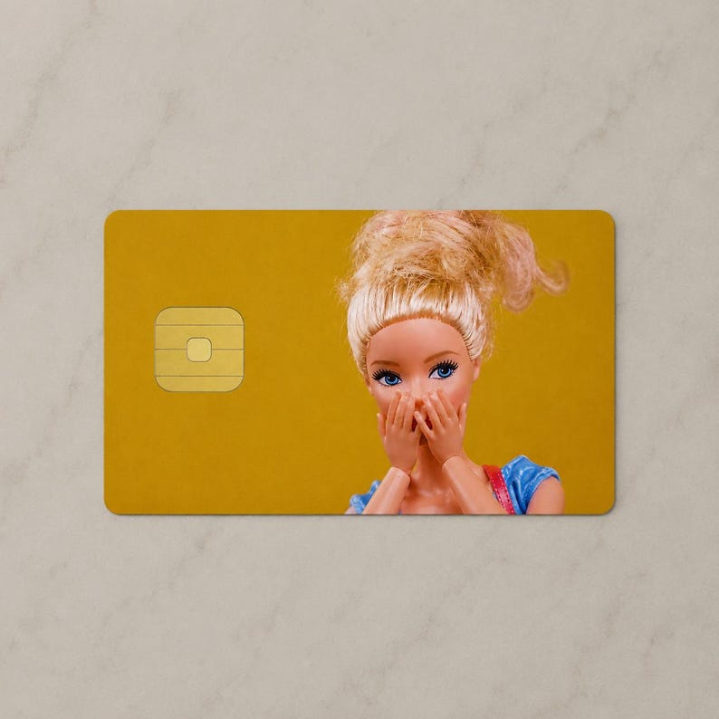 Pu&ograve; includere: Una carta di credito rettangolare con sfondo giallo senape, un chip EMV dorato sul lato sinistro e una bambola con capelli biondi raccolti in uno chignon, occhi azzurri e mani che coprono la bocca sul lato destro.