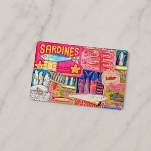 May include: A rectangular, colorful illustration featuring various sardine tins and fish imagery. The design incorporates pink, blue, yellow, and red hues, with the word "SARDINES" prominently displayed. The artwork has a vintage, hand-drawn aesthetic.