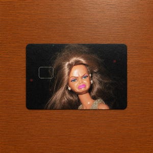 May include: A rectangular credit card skin featuring a close-up of a doll with long brown hair, a furrowed brow, and bright pink lips. The doll is set against a solid black background, and the card includes a cutout for a chip reader.