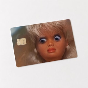 Op de afbeelding: Een rechthoekige kaart met een poppengezicht met grote, blauwe ogen en een verbaasde uitdrukking. De pop heeft blond haar en een roze-rode lipkleur. Een goudkleurige chip bevindt zich in de linkerbovenhoek van de kaart.