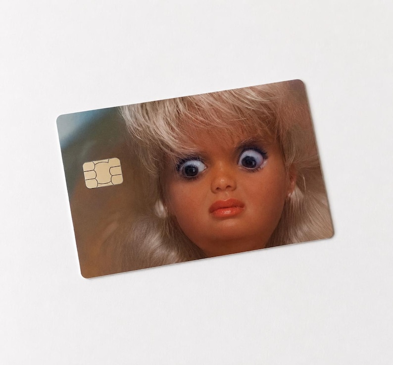 Op de afbeelding: Een rechthoekige kaart met een poppengezicht met grote, blauwe ogen en een verbaasde uitdrukking. De pop heeft blond haar en een roze-rode lipkleur. Een goudkleurige chip bevindt zich in de linkerbovenhoek van de kaart.