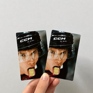 Op de afbeelding: Twee rechthoekige kaarten met een hockeyspeler met een CCM-helm met een helder vizier. De tong van de speler steekt uit en houdt een simkaart vast. De kaarten worden door een hand vastgehouden.