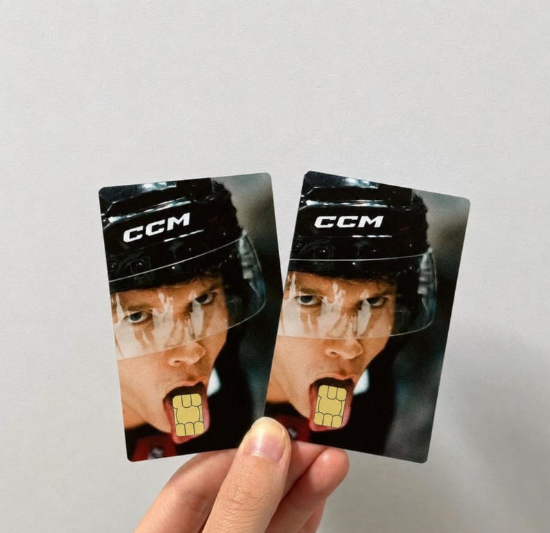 Op de afbeelding: Twee rechthoekige kaarten met een hockeyspeler met een CCM-helm met een helder vizier. De tong van de speler steekt uit en houdt een simkaart vast. De kaarten worden door een hand vastgehouden.