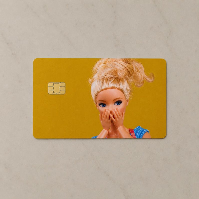 Pu&ograve; includere: Una carta di credito rettangolare con uno sfondo giallo senape solido e un chip metallico dorato sul lato sinistro. Il lato destro mostra una bambola con capelli biondi raccolti in uno chignon spettinato, occhi blu e mani sulla bocca.