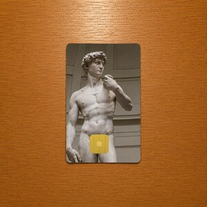 Op de afbeelding: Een rechthoekige kaart met een grijswaardenafbeelding van het David-beeld van Michelangelo. Het naakte beeld heeft een gele vierkant die de geslachtsdelen bedekt. De kaart staat tegen een houten achtergrond.
