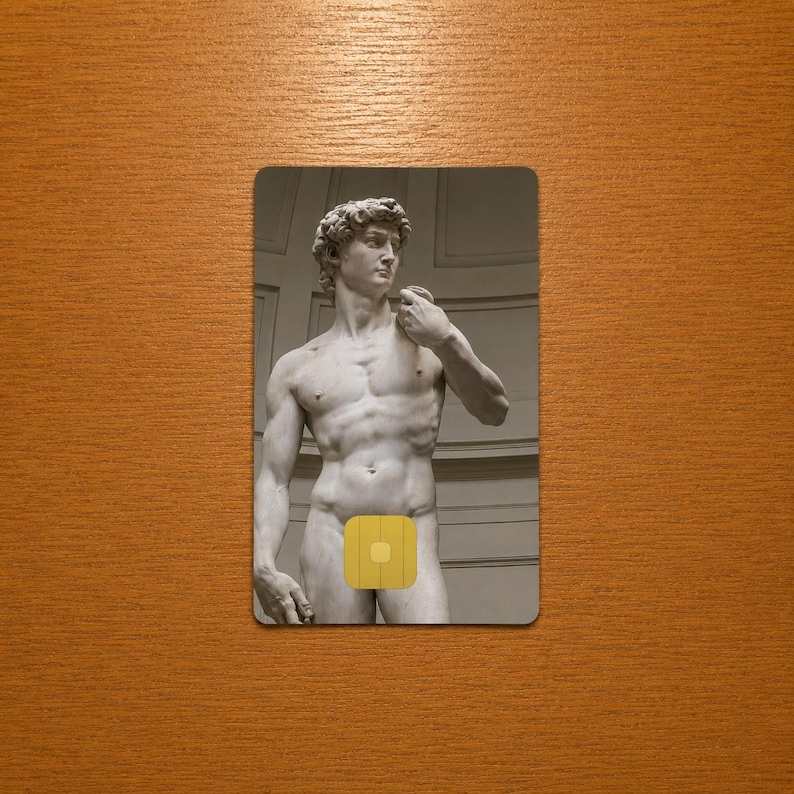Op de afbeelding: Een rechthoekige kaart met een grijswaardenafbeelding van het David-beeld van Michelangelo. Het naakte beeld heeft een gele vierkant die de geslachtsdelen bedekt. De kaart staat tegen een houten achtergrond.
