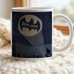 Puede incluir: Una taza de cerámica blanca con un gráfico de cielo nocturno azul oscuro y un foco en forma de murciélago que ilumina una ciudad. Dentro del foco, el texto dice BRING COFFEE sobre una pequeña silueta de una figura en una cornisa.