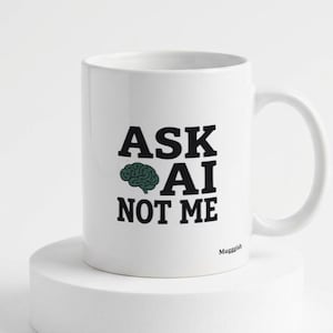 Könnte beinhalten: Ein weißer Keramikbecher mit dem schwarzen Text ASK AI NOT ME und einem kleinen grünen Gehirnsymbol zwischen den Wörtern ASK und AI. Das Wort Muggish ist in kleinen schwarzen Buchstaben unten rechts auf dem Becherdesign aufgedruckt.