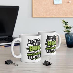 May include: Two white ceramic mugs featuring a black and green graphic design with the text "I DON'T HAVE A BUG I HAVE AN UNDOCUMENTED FEATURE" and a small cartoon insect icon. The mugs have a standard cylindrical shape with a curved handle.