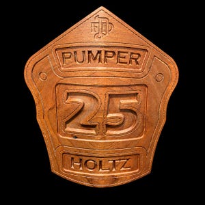 May include: A shield-shaped wooden plaque with a natural brown finish. The top features an engraved emblem, followed by the word PUMPER, the large number 25 in the center, and the name HOLTZ at the bottom, all with carved lettering.