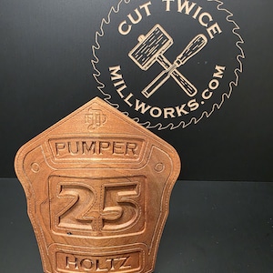 May include: A wooden firefighter badge with the words "PUMPER 25 HOLTZ" carved into it. The badge is a warm brown color and has a shield shape. In the background is a circular logo with the text "CUT TWICE MILLWORKS.COM".