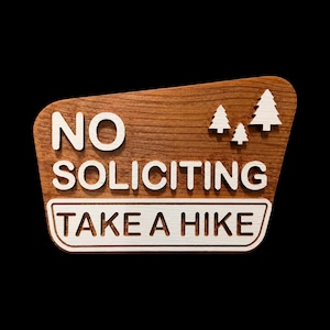 Könnte beinhalten: Ein Holzschild mit natürlicher Holzmaserung und weißer, erhabener Schrift mit der Aufschrift NO SOLICITING und TAKE A HIKE. Das Schild enthält drei weiße, ausgeschnittene Tannenbaumformen, die rechts über dem Text angebracht sind.