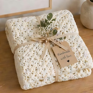 May include: A folded, cream-coloured crocheted blanket tied with a beige ribbon and a sprig of greenery. A small tag reads "Handmade with love" with a heart symbol. The blanket is presented as a gift.