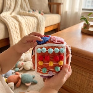 May include: A crocheted sensory cube toy featuring a rust-orange base with white borders. One side displays an abacus-style design with four horizontal rows of small, round, multi-colored crocheted beads in light blue, red, yellow, and teal.