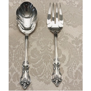 May include: A silver-toned serving spoon and a matching four-tined serving fork. Both utensils feature a polished, reflective finish and intricate, embossed floral detailing along the handles, which taper into a decorative, ornate base design.