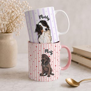 以下が含まれることがあります： 犬のイラストが描かれた2つの積み重ねられたセラミックマグ。上のマグは白いハンドルで、紫色のストライプ、ハート、そして「Marley」という名前で装飾されています。下のマグはピンクのハンドル、ピンクのストライプ、赤いハート、そして「Molly」という名前です。
