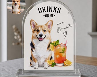 May include: A clear acrylic sign with a watercolor illustration of a Corgi dog and a cocktail. The sign reads "DRINKS ON ME" with the name "Barnie" and a heart. The cocktail has a straw, ice, and a lemon slice.