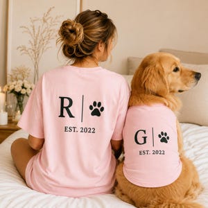 May include: Matching pink t-shirts with black text and paw prints. One shirt has the letter "R" and the other has the letter "G", both with "EST. 2022". A golden retriever dog wears a matching shirt.