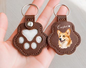 May include: Two brown leather paw-shaped keychains with silver metal rings. One keychain features a cutout paw print design with white fur inside. The other keychain displays a portrait of a Corgi dog with the name Casper and a heart printed above.