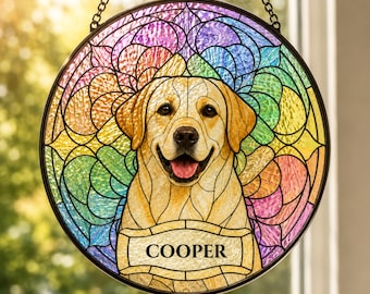 May include: A circular stained glass suncatcher featuring a yellow Labrador retriever with an open mouth and pink tongue. The background consists of a multi-colored, rainbow-toned geometric pattern. A rectangular plaque at the bottom reads COOPER.