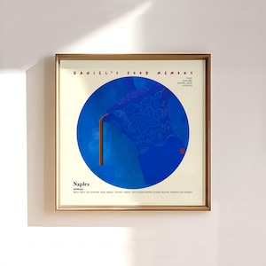 Op de afbeelding: Een ingelijste kunstdruk met een blauw en bruin grafisch ontwerp. Het kunstwerk bevat een grote blauwe cirkel met een gedetailleerd patroon en een bruin element. De tekst onderaan luidt "Naples Espresso". De lijst is goudkleurig.