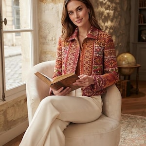 May include: A long-sleeved jacket featuring a vibrant, multi-coloured tapestry-style pattern in shades of red, orange, and gold. The jacket has a structured collar, a full-length front zip, and a fitted silhouette, paired with solid cream trousers.
