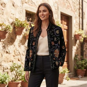 May include: A black velvet jacket featuring long sleeves and a stand-up collar, adorned with intricate floral embroidery in shades of orange, blue, and green. The jacket is worn over a cream-coloured camisole and paired with black trousers.