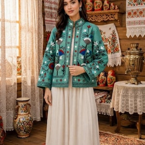 Può includere: Una giacca color verde acqua con colletto alla coreana e maniche lunghe, decorata con intricati ricami floreali in rosso, bianco e blu. La giacca è indossata sopra un lungo abito color crema con una sottile trama verticale e una linea fluida.
