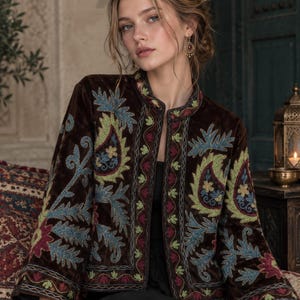 May include: A dark brown velvet jacket featuring an open front and a mandarin collar. The garment is adorned with intricate, multi-colored embroidery in shades of light green, blue, and deep red, showcasing floral and paisley-inspired patterns.