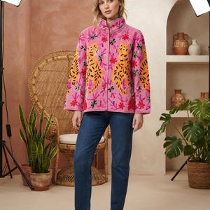 Könnte beinhalten: Eine rosafarbene Langarmjacke mit gestickten gelben und schwarzen Leoparden, umgeben von bunten Blumenmustern, kombiniert mit einer dunkelblauen Jeans und weißen Turnschuhen. Die Jacke hat einen Stehkragen und eine Knopfleiste.